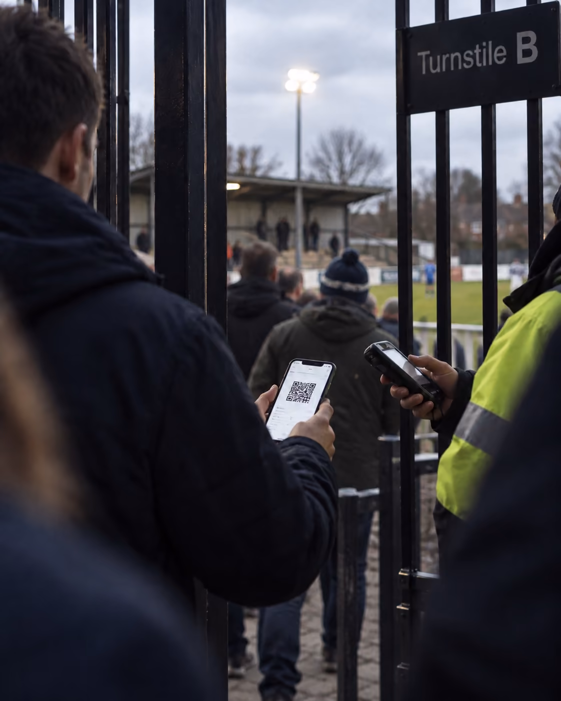 A fan showing the QR ticket on their phone to a steward with a scanner at the turnstile of a small football ground.